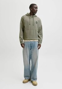 Vihreä huppari edustataskulla ja logolla, yhdistettynä väljään, vaaleansinisiin farkkuihin ja beigenvärisiin kenkiin. Sileä kangas ja rento istuvuus.