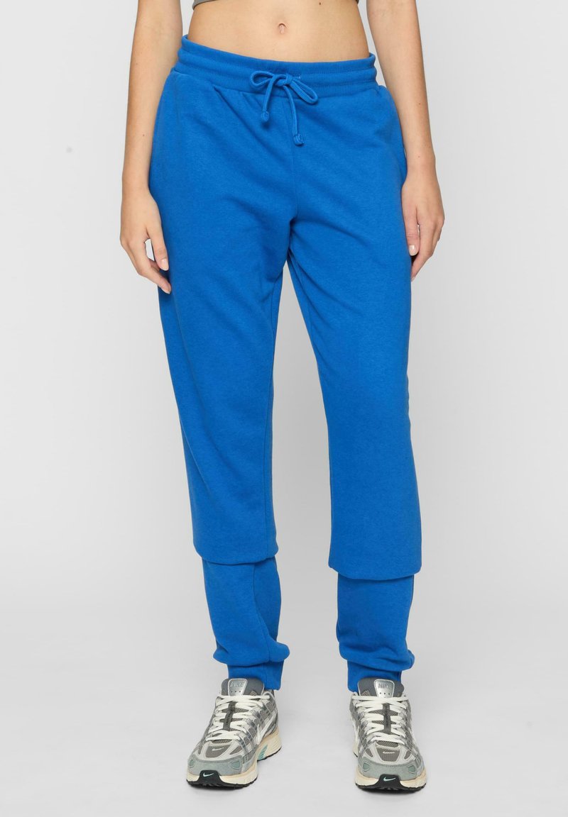Blauwe sweatpants met elastische tailleband en koord. Zacht katoenen materiaal. Omgeslagen onderkanten. Draag met grijze en zilveren sneakers.