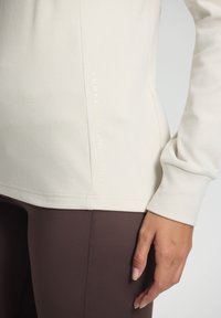 Close-up of a person’s hand resting by their side wearing a beige long-sleeve top with "Luhta Outdoor Lifestyle" text and brown leggings.