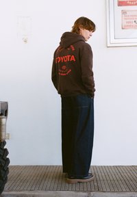 Hnedá mikina s kapucňou s červeným nápisom "TOYOTA" na chrbte, doplnená voľnými tmavými denimovými nohavicami a hnedou obuvou. Prostredie má textúrovanú podlahu.