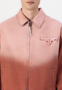 Pink denim jacket with a zipper front, pointed collar, chest pocket featuring triangular detail, and a smooth texture with a gradient effect.