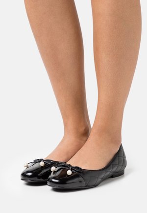 Des pieds chaussés de ballerines noires matelassées avec des nœuds et des perles en suspension sur un fond blanc.