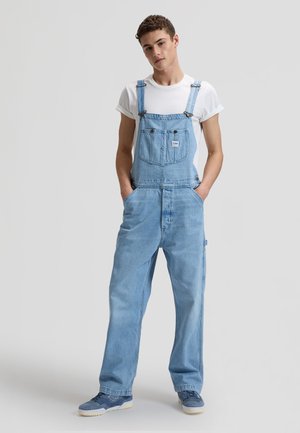 Junger Mann steht mit den Händen in den Taschen, trägt hellblaue Jeans-Latzhose, weißes T-Shirt und blaue Sneaker vor einfachem Hintergrund.