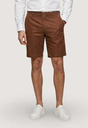 Homme portant un short marron sur mesure jusqu'aux genoux, une chemise blanche boutonnée et des baskets blanches basses, debout avec une main dans la poche.