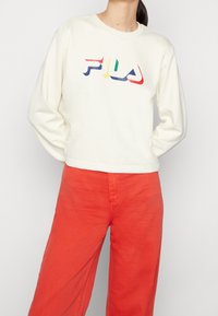 Cremfarbener Sweatshirt mit langen Ärmeln und Rundhalsausschnitt, versehen mit einem mehrfarbigen Logo. Kombiniert mit hochwaisted, leuchtend orangefarbenen Hosen.