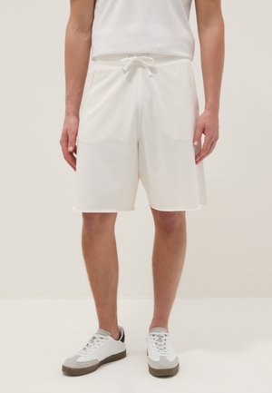 Person wearing white drawstring shorts and white sneakers with gray accents, standing against a light neutral background.