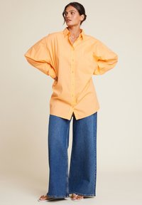 Overdimensjonert oransje bomullsskjorte med lange ermer, en frontlomme og knappelukking, kombinert med vide blå denimjeans.