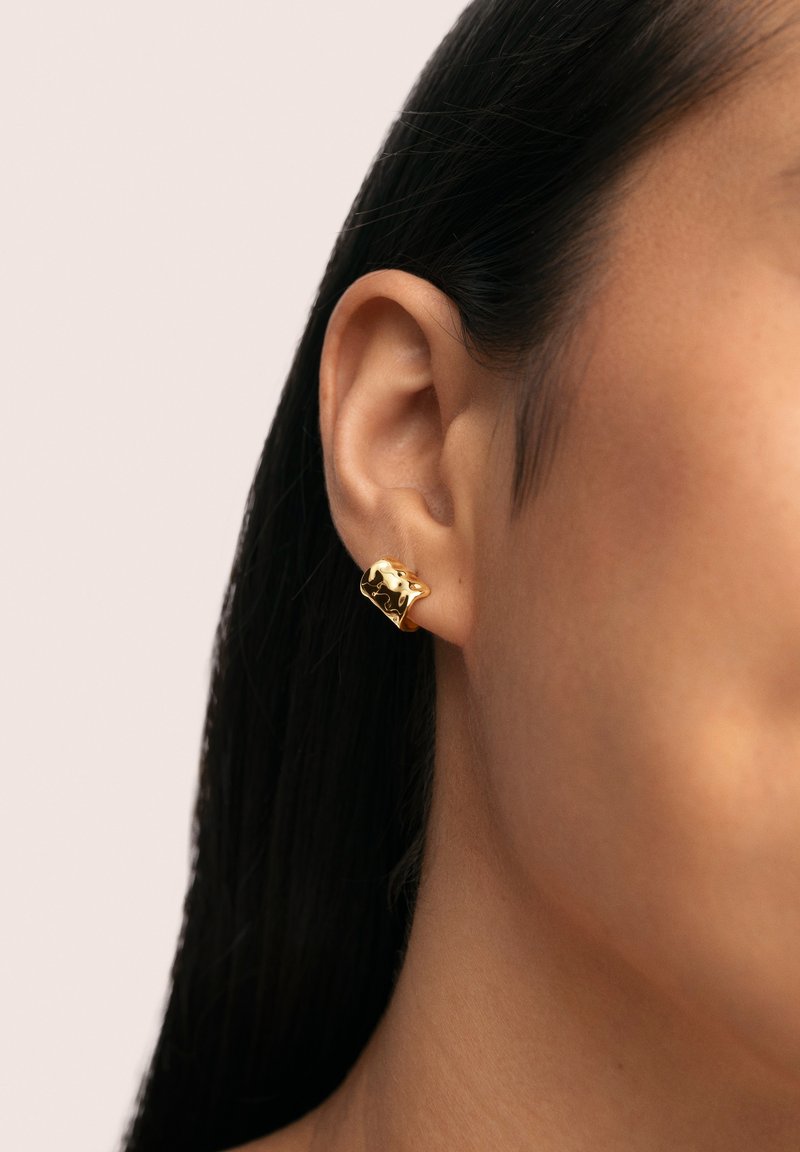 Goldfarbene Metallohrringe mit einem abstrakten Design. Der Ohrring ist klobig und glatt und ist am Ohr einer Person mit glattem schwarzen Haar befestigt.