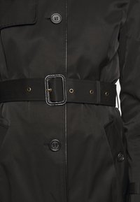 Manteau imperméable noir avec une taille ceinturée, deux poches avant et une fermeture à boutons. Texture lisse avec des accents de ferrures dorées.