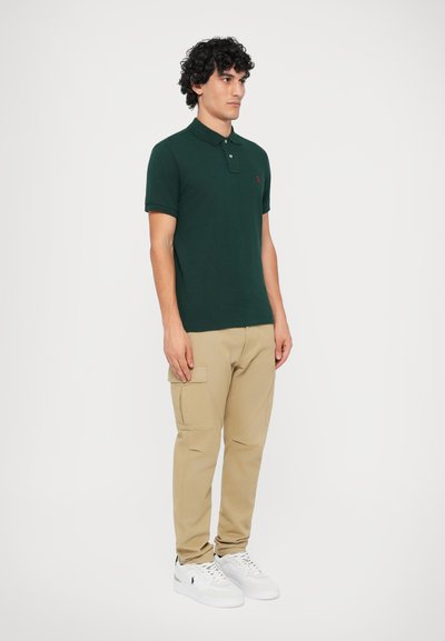 Grön pikétröja med krage och röd emblem, tillsammans med beige cargobyxor med sidofickor, och vita sneakers.