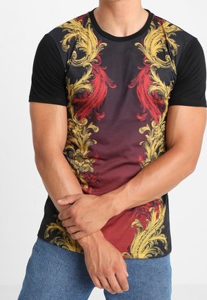 Uomo che indossa una maglietta nera a maniche corte con motivi floreali ornamentali rossi e dorati, abbinata a jeans blu, braccia incrociate in vita.