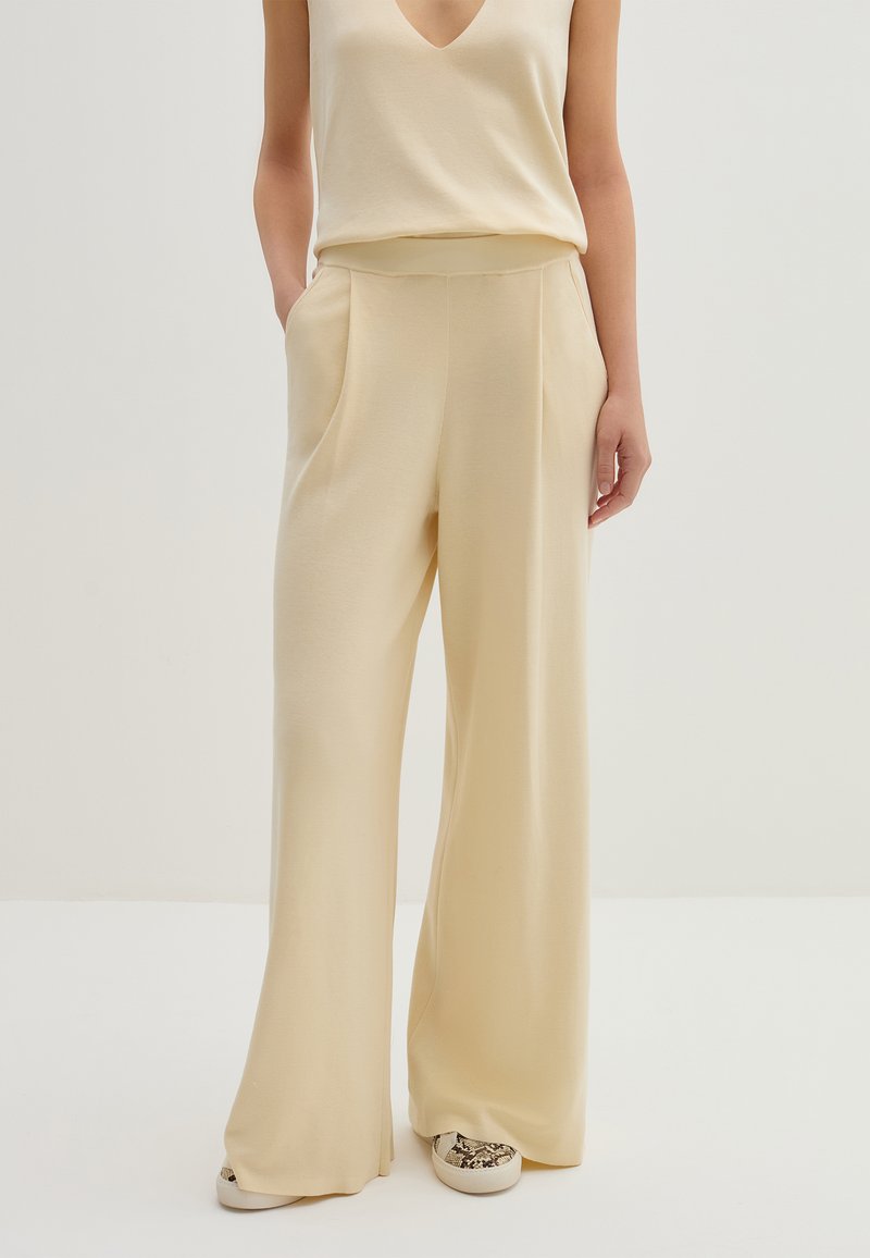Pantaloni beige a gamba larga con texture liscia, dotati di tasche laterali e vita alta, abbinati a un top senza maniche coordinato.