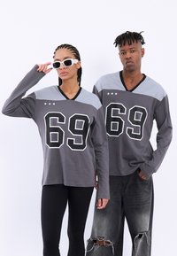 Grå langermede skjorter med svart og hvit '69'-trykk og v-halsdesign, kombinert med svarte ribbede leggings og slitte grå jeans.