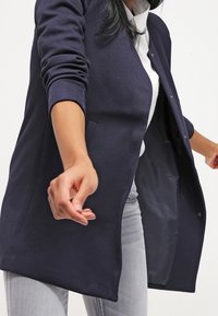 Mörkblå långärmad kavaj med texturerad tyg, öppen front och två sidofickor, bärs över en vit skjorta och ljusgrå jeans.