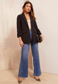 Musta bleiseri, jossa on pitkät hihat ja etutaskut, yhdistettynä korkeavyötäröisiin levennettyihin sinisiin farkkuihin ja leopardikuvioisiin korkokenkiin. Minimalistinen musta clutch-laukku.