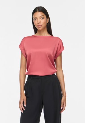 Femme aux longs cheveux foncés portant une blouse corail à manches retroussées rentrée dans un pantalon noir taille haute, debout sur fond blanc.