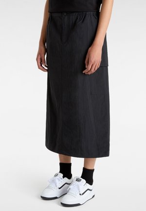 Person trägt einen langen schwarzen Rock mit Taschen, schwarze Socken und weiße Vans Sneakers und steht vor einem einfarbigen Hintergrund.