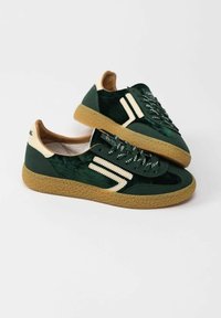 Sneakers verde in suede con suola in gomma testurizzata, accenti crema e strisce laterali a contrasto. Presentano la chiusura con lacci e linguetta imbottita.