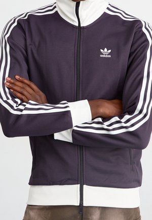 Osoba ubrana w ciemnofioletową kurtkę sportową Adidas z białymi paskami na rękawach, białymi mankietami i suwakiem z przodu, z założonymi na piersi złożonymi ramionami.