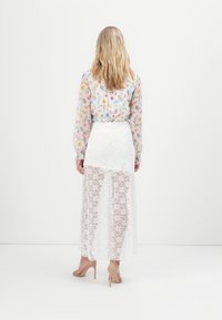 Blusa translúcida con estampado floral y mangas largas, combinada con una falda larga de encaje blanca, que presenta un diseño en capas y un sutil patrón floral.
