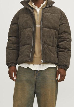Homme portant une doudoune marron en velours côtelé sur un pull beige et une chemise blanche, associée à un jean ample délavé.