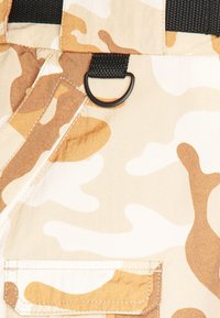 Tissu camouflage beige et marron avec sangle noire, anneau en D, couture détaillée et rabat de poche.