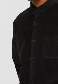 Homme portant une chemise noire en velours côtelé à boutons avec poche poitrine et col, sur un fond uni gris clair.