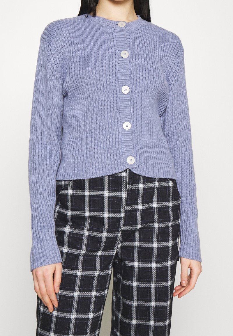 Femme portant un cardigan boutonné côtelé violet clair et un pantalon à carreaux noir et blanc, debout devant un fond blanc uni.