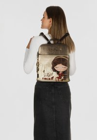 Der Rucksack verfügt über eine metallisch goldene Oberseite, ein illustriertes Design mit einem Mädchen in Rot und den Text "KID YOURSELF." Die Stofftextur ist glatt mit stabilen Trägern.