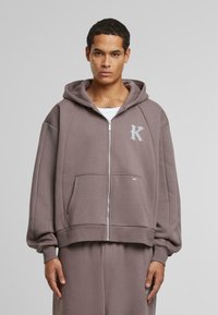 Felpa con cappuccio e zip in marrone tenue, con logo sul petto, tasche frontali e polsini a costine, dal fit rilassato.