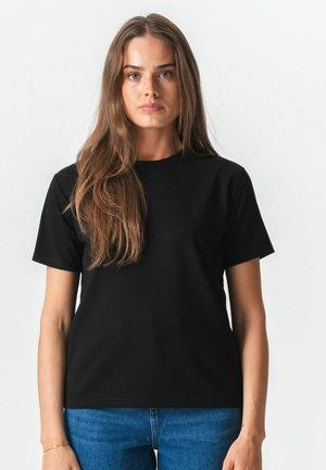 Kvinde med langt brunt hår iført en ensfarvet sort t-shirt med korte ærmer og blå jeans, stående foran en hvid baggrund.