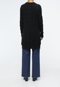 Sort strikket lang cardigan med tekstureret bladmønster, brede ærmer og ribbede manchetter, parret med tætsiddende blå jeans og sorte chunky hæle.