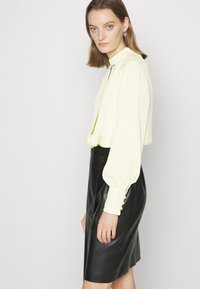 Blusa amarelo claro com mangas longas e bufantes e punhos abotoados, combinada com uma saia justa de couro preto. Design elegante e moderno.