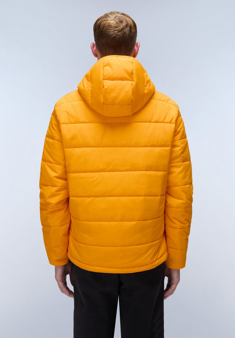 Uomo che indossa un giubbotto puffer arancione brillante con cappuccio, in piedi con la schiena rivolta verso uno sfondo grigio chiaro uniforme.