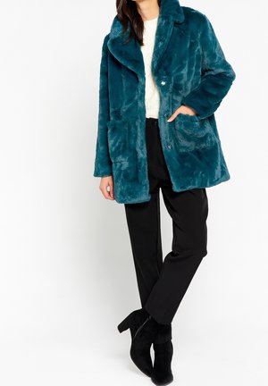 Kobieta w turkusowym futrzanym płaszczu z imitacji futra, białym swetrze, czarnych spodniach i czarnych zamszowych botkach na białym tle.