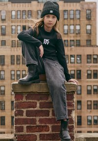 Joven con un gorro negro, suéter negro de DKNY, jeans grises y botas negras sentado en un borde de ladrillo con edificios urbanos de fondo.