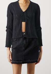 Cardigan nero lavorato a maglia con scollatura a V e chiusura con lacci, caratterizzato da un design a trafori. Abbinato a una gonna in denim scuro con orlo sfilacciato.