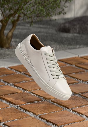 Weißer Leder-Sneaker mit hellgrauen Schnürsenkeln, der über gemusterten Holzkacheln mit kleinen grauen Steinen dazwischen schwebt.