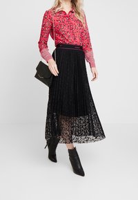 Rød leopardmønstret bluse med lange ærmer, parret med en sort plisseret blonde-nederdel. Tilbehør inkluderer en lille sort håndtaske og ankelstøvler.