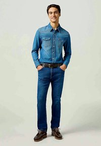 Camisa de mezclilla azul con dos bolsillos en el pecho y mangas largas, combinada con jeans de mezclilla azul y zapatos marrones, llevada por una figura masculina.