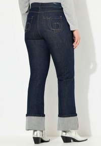 Jeans de mezclilla de lavado oscuro, acampanados y con dobladillo. Presenta dos bolsillos traseros con un diseño cosido y costuras en contraste.