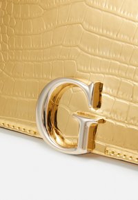 Guldkrokodillembossed materiale med en blank overflade, der har et fremtrædende sølvfarvet 'G' hardwaredetalje. Fint synlige stikninger.