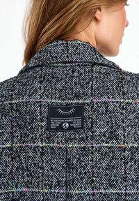 Žena v čierno-bielom kabáte s rybím vzorom a farebnými károvanými líniami, s nášivkou na hornej časti chrbta nesúcou poetickú správu.