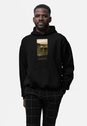 Mann trägt schwarzen Hoodie mit Pferderennen-Grafik und dem Text "Edgar Degas Longchamp, Frankreich" sowie schwarze Hose mit weißem Karomuster.