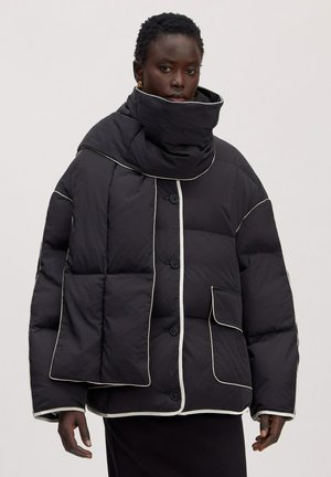 Schwarze übergroße wattierte Jacke mit kontrastierenden weißen Paspeln, getragen von einem Model mit kurzen Haaren vor einfarbigem Hintergrund.