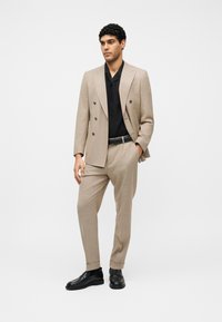 Homme debout, une main dans la poche, portant un costume beige croisé, une chemise noire, une ceinture noire et des chaussures de ville noires sur un fond uni.