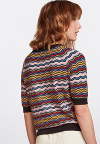 Sweter dzianinowy z wielokolorowym wzorem w zygzaku w odcieniach różu, żółci, zieleni i czerwieni. Krótkie rękawy i ciemny ściągacz u dołu.