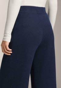 Tmavomodré rebrované pletené nohavice so širokými nohavicami, elastickým pásom a textúrovaným vzorom. Pohľad zozadu, s rukou položenou na boku.