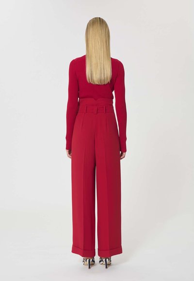 Femme aux longs cheveux blonds vue de dos, portant un haut rouge à manches longues, un pantalon rouge taille haute à jambes larges, et des escarpins à motifs.
