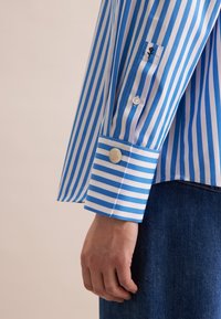 Camicia a righe blu e bianche con bottoni, maniche lunghe, polsini con bottone e un dettaglio ricamato sulla manica.